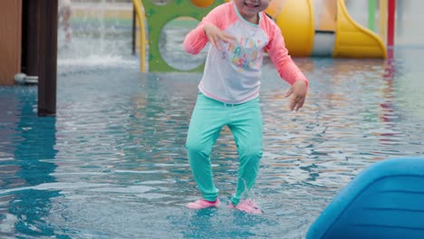 excited little girl splashing water with hands and jumps playing at outdoor water playground during summer holidays