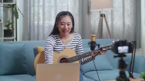 asian woman reading comments on laptop and speaking to camera while live stream playing a guitar at home