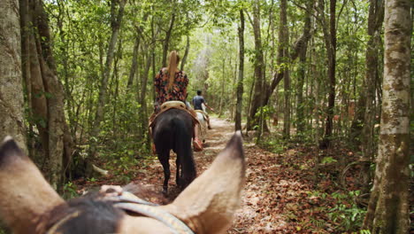 slow motion back shot of young blonde girl with flower dress horseback riding inside tropical forest pathway from horse prospective in cancun, mexico