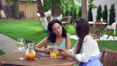 two women sitting at the outdoors cafe talking, sharing news, girls showing something on laptop screen and smartphone