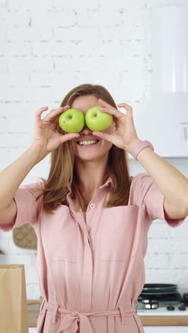 smiling girl in the kitchen holds green apples in her hands and closes her eyes with them. healthy eating concept. woman on a diet. vertical video