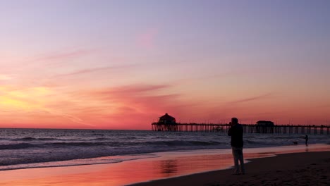 a man takes a picture of the beach during a gorgeous yellow, orange, pink and blue sunset with the huntington beach pier in the background at surf city usa california