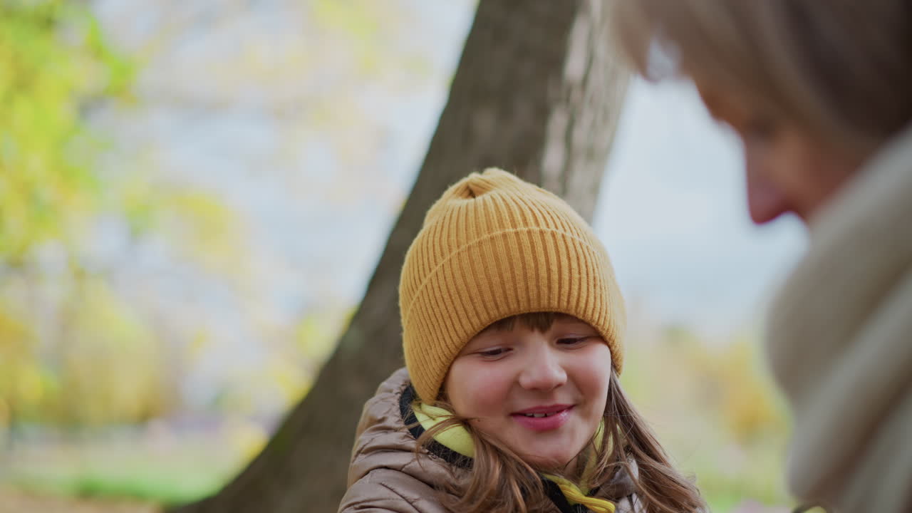 little girl stands behind mother in autumn park waving golden leaves to get mother attention while mother turns away on bench engrossed in work on laptop amidst colorful scenery of trees and grass
