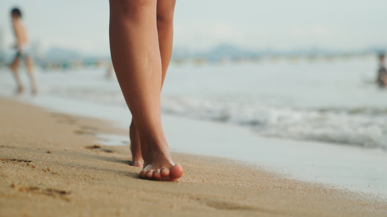 Piedi che camminano lungo la riva sabbiosa, onde leggere che lambiscono le caviglie, viaggiatore solitario che passeggia oltre nuotatori lontani, calda luce solare che proietta lunghe ombre, impronte che si formano e poi vengono cancellate dalla marea, lento.