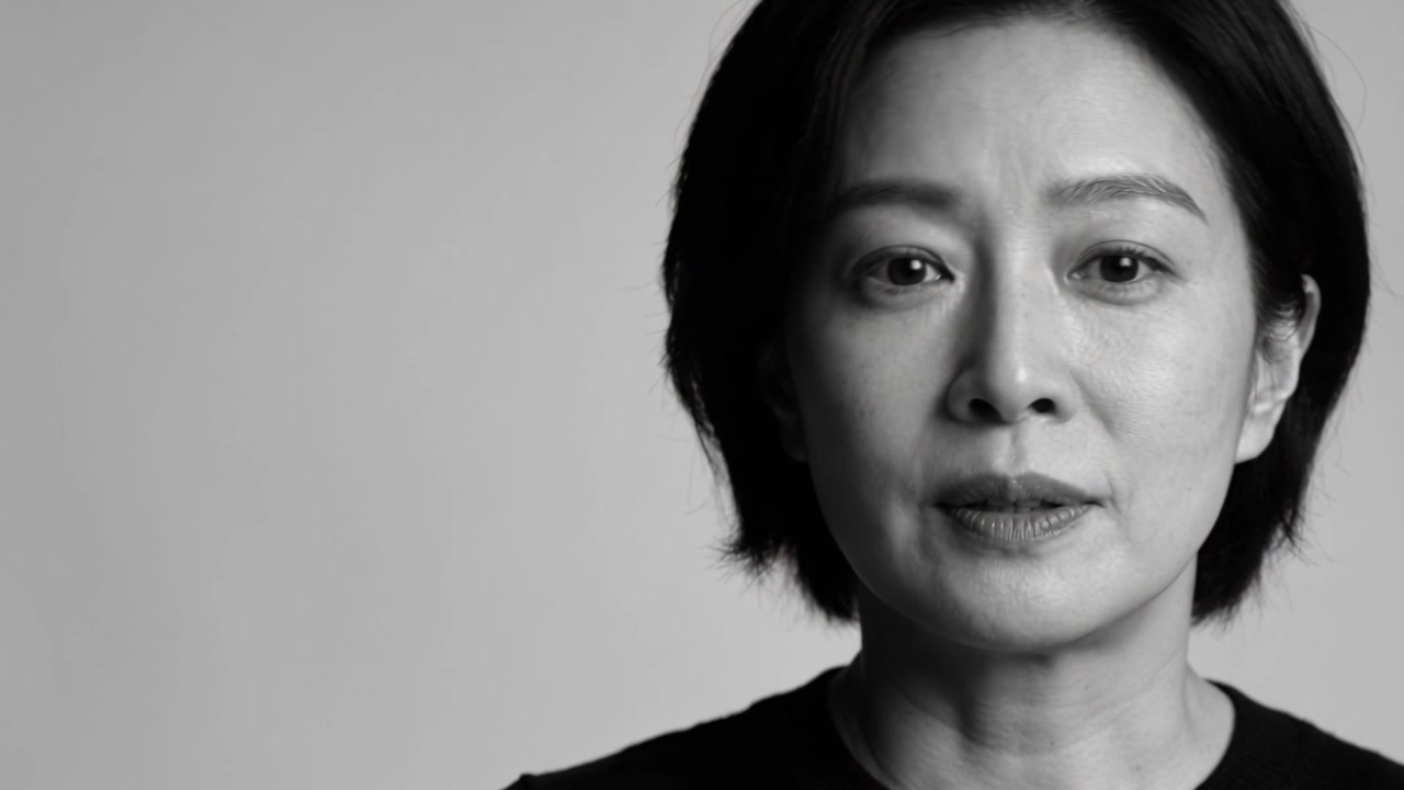 Turning Asian woman speaking to camera in studio, giving speech, plain backdrop, wearing black top