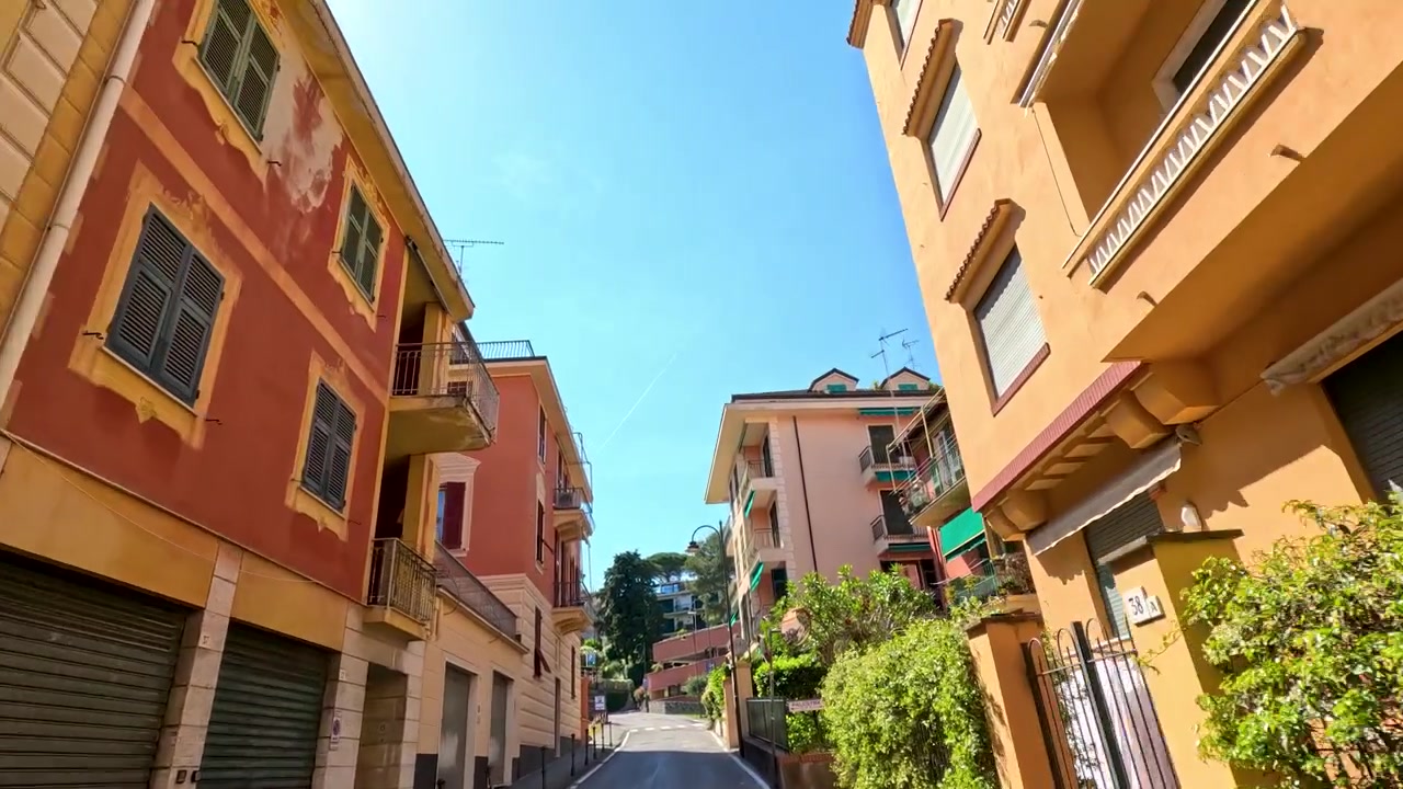 Carrellata dal basso che si muove attraverso una strada stretta con edifici color pastello e finestre con persiane chiuse.