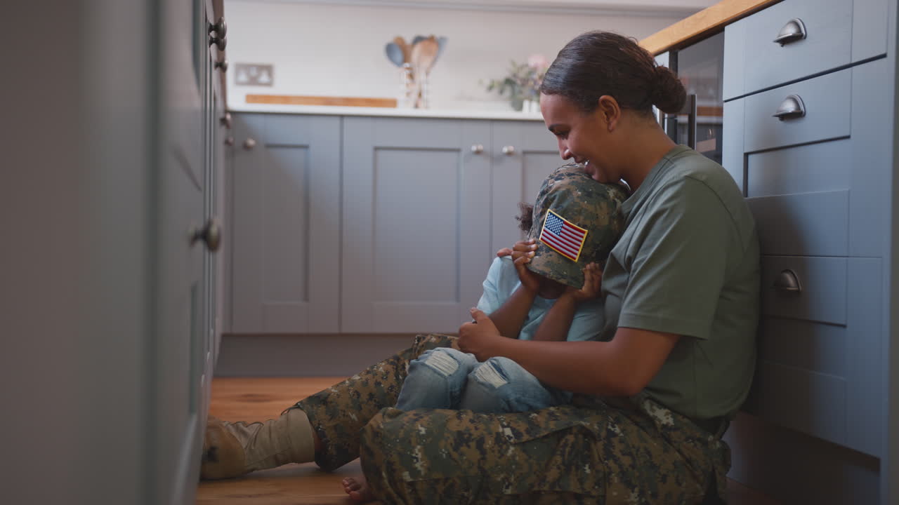 madre del ejército estadounidense en uniforme en casa de licencia abrazando a su hijo sentado en el suelo en la cocina de la familia