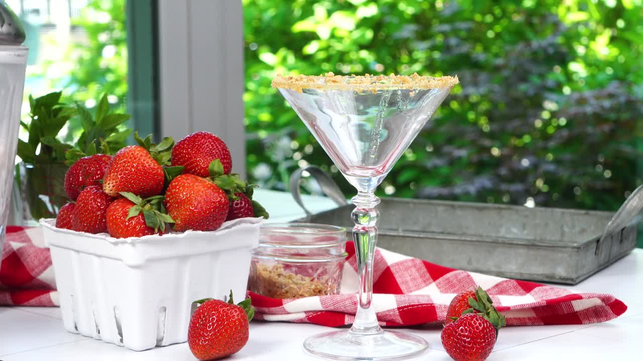 Strawberry Shortcake Martini cocktail with whipped cream, graham cracker crumbs and a fresh red strawberry. Pouring the cocktail into the martini glass. A summer time berry drink with red napkin.