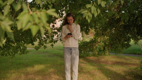lady in casual clothing stands under tree, deeply focused on phone with hand on hood collar, sunlight filtering through leaves, with gentle greenery in background swaying slightly