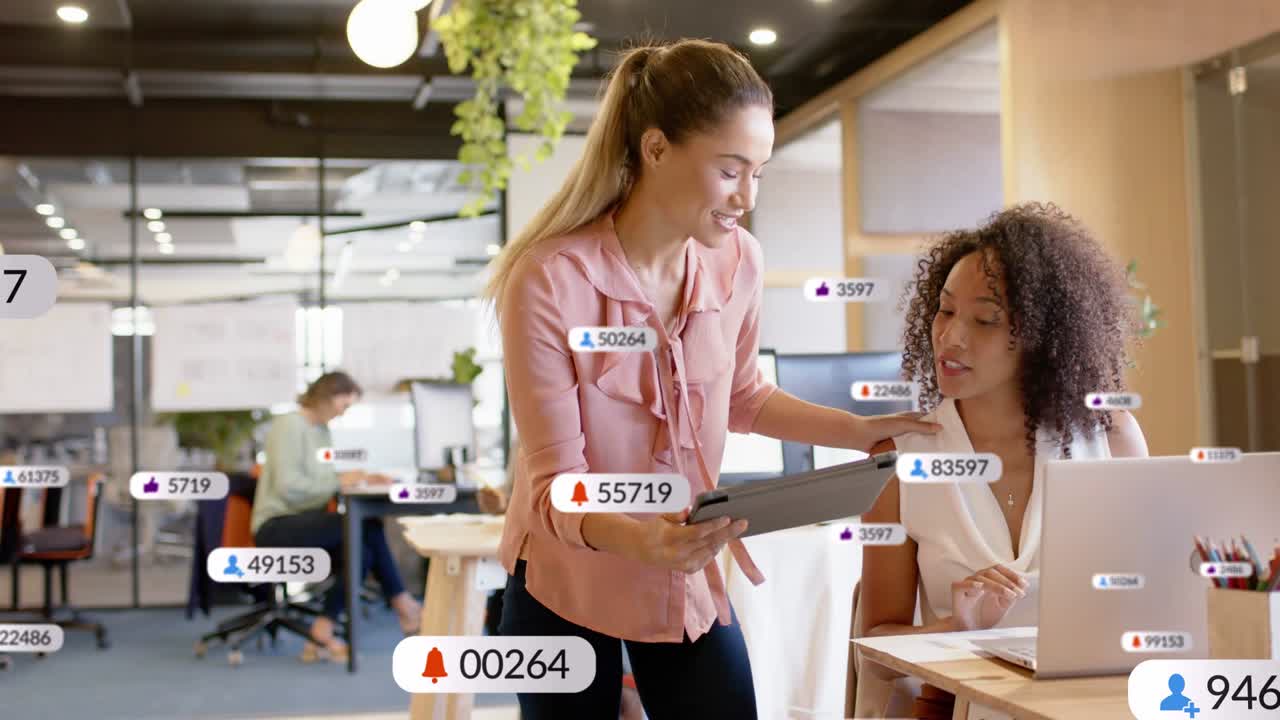 Standing woman showing tablet seated peer reviewing metrics on laptop badges rising across business
