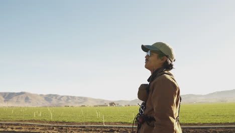 Medium-shot-of-a-man-with-a-heavy-leather-glove-hear,-thick-rim-gasses-and-mustache-searching-the-sky-near-a-green-farm-field-surrounded-by-dry-brown-mountains