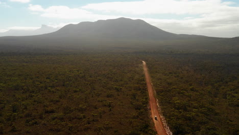 Aerial-view-overlooking-a-4x4-vehicle,-driving-on-a-red-sand,-desert-road,-towards-a-huge-mountain,-bright,-sunny-day,-in-Hassell-National-Park,-West-Australia---descending,-drone-shot