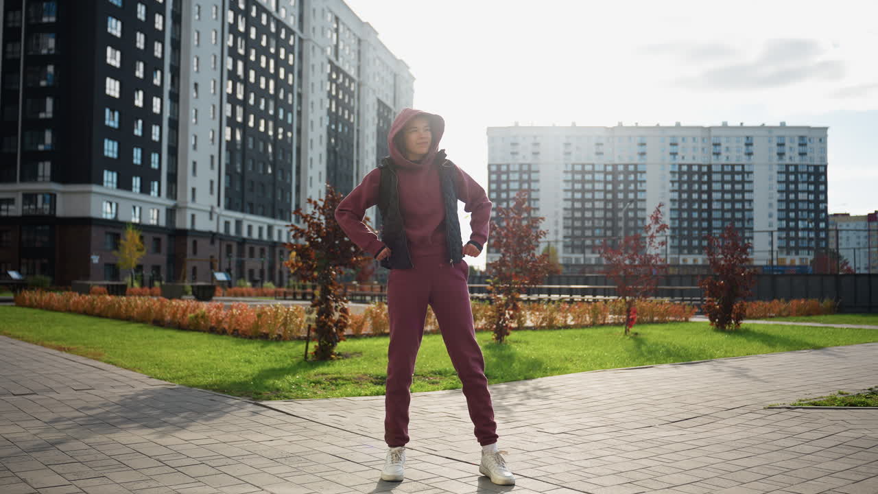 Trainer wearing mauve hoodie and black vest jacket performs side stretch under sunny sky on urban paved plaza amid high rise buildings and autumn foliage during outdoor exercise session