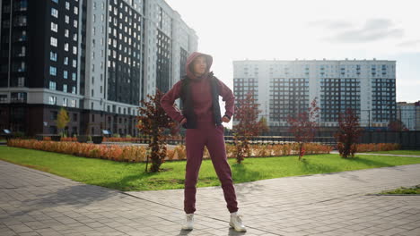 trainer wearing mauve hoodie and black vest jacket performs side stretch under sunny sky on urban paved plaza amid high rise buildings and autumn foliage during outdoor exercise session