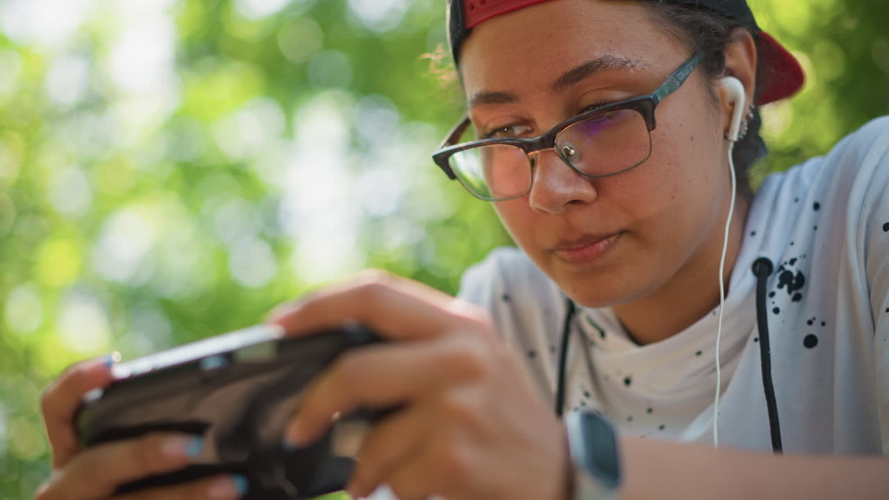 공원에서 촬영한 아시아인 게이머의 인물 사진, 안도하는 표정과 부드러운 미소, 집중하는 시선, 이어폰 착용, 안경 착용, 휴대폰을 안정적으로 잡은 손, 나뭇잎 사이로 부드럽게 비치는 햇빛, 미묘한 감정을 포착한 클로즈업 사진