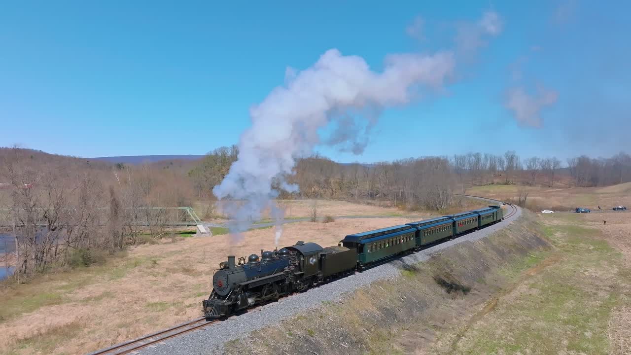 A steam locomotive chugs along winding tracks, releasing puffs of smoke as it passes through open fields and distant hills under a bright blue sky.