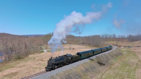 a steam locomotive chugs along winding tracks, releasing puffs of smoke as it passes through open fields and distant hills under a bright blue sky.