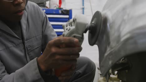 African-American-male-car-mechanic-using-an-angle-grinder-on-the-side-of-a-car-