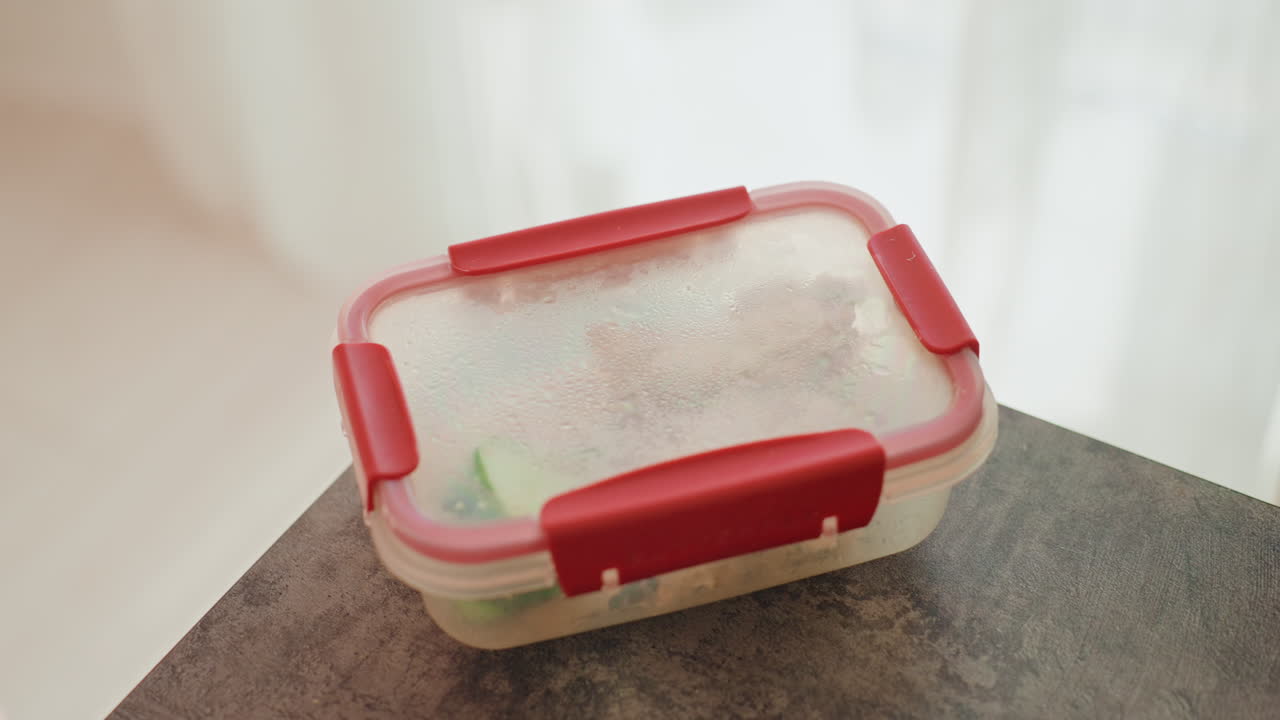 Woman Prepares Meal, Female Individual Revealing Food Inside Lunchbox With Peeled Lid, Woman In Kitchen Carefully Opening Lunchbox Revealing Cooked Chicken And Fresh Cucumber Ready To Serve