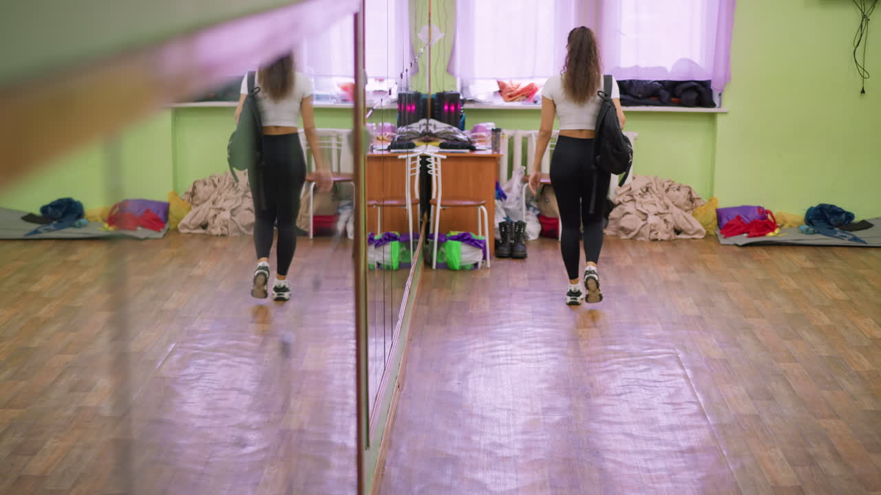 Lady in white top and black leggings carrying school bag closes door and walks toward window, body reflecting in large wall mirror inside bright indoor room with wooden floor and soft daylight