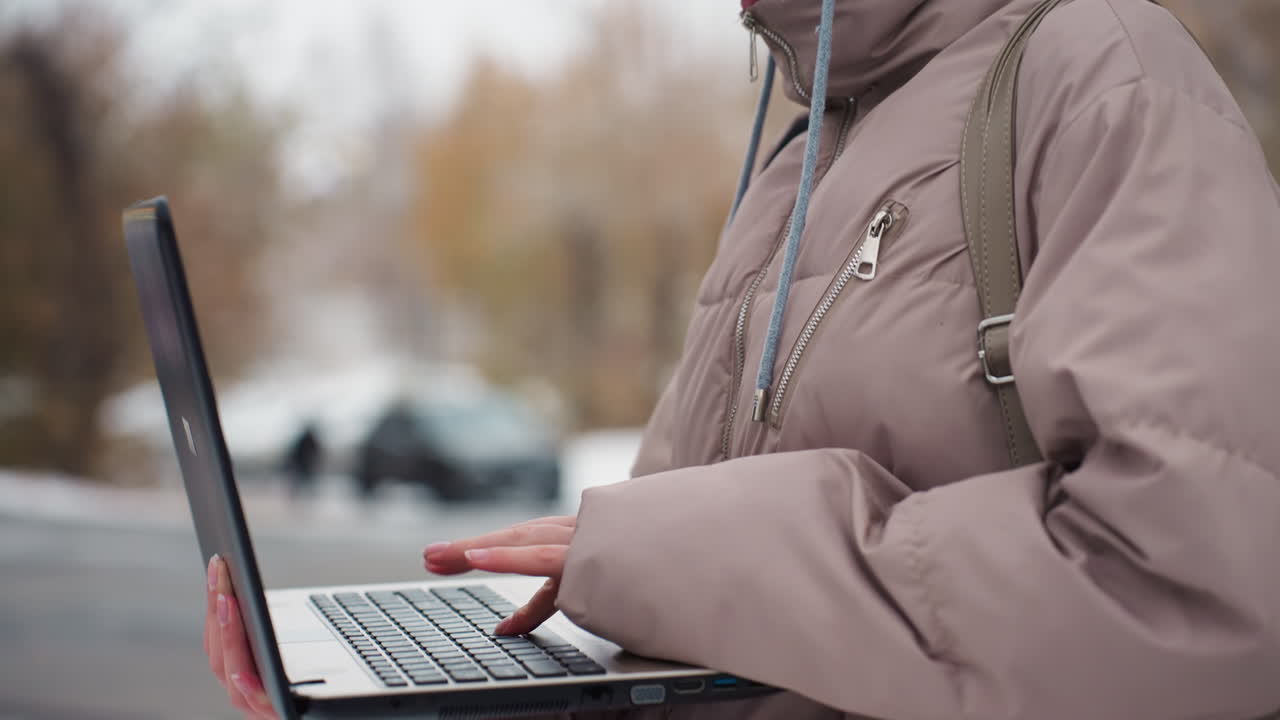 Side view of light skin lady in beige beanie and brown winter jacket concentrating while operating laptop outdoors, with soft blur background of autumn trees and distant people