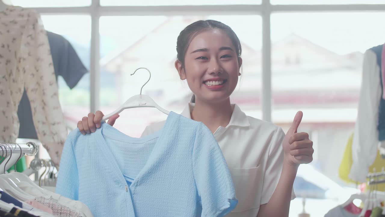 젊은 아시아 여성이 파란색 옷을 선택하고 미소를 지으며 부티크에서 쇼핑하는 동안 카메라에 엄지손가락을 보여주고 있습니다.