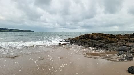 slow motion calming ocean waves washing across anglesey island splashing over rocks on golden sandy beach, overcast morning