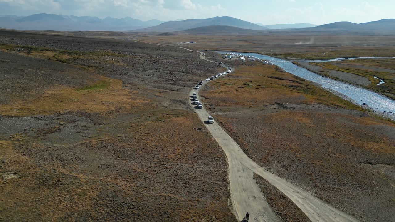 vista aérea de un convoy de vehículos que conducen por una pista de tierra a lo largo de un río en un vasto paisaje abierto, mostrando un sentido de aventura y exploración