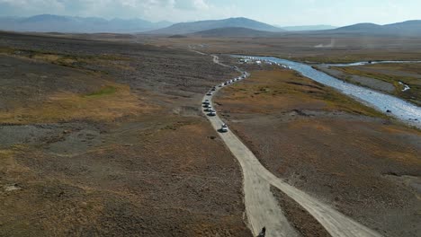 Luftaufnahme-Eines-Konvois-Von-Fahrzeugen,-Die-Auf-Einem-Feldweg-Entlang-Eines-Flusses-In-Einer-Weiten,-Offenen-Landschaft-Fahren-Und-Ein-Gefühl-Von-Abenteuer-Und-Entdeckung-Vermitteln