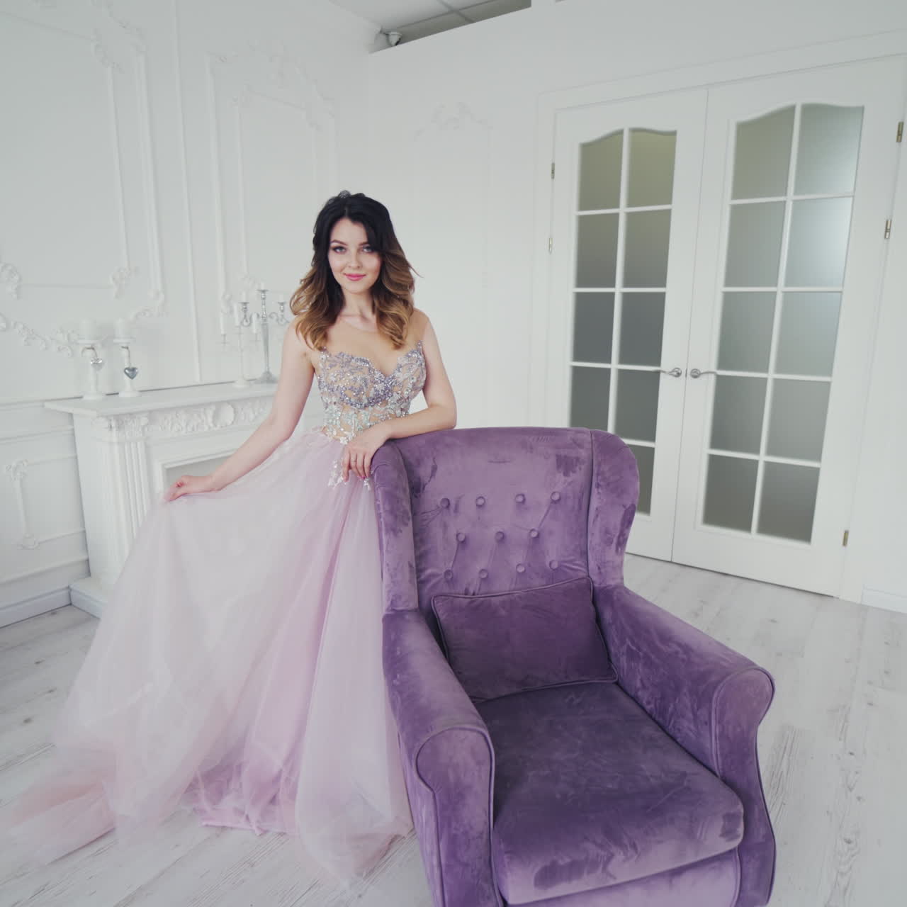 Fantastic young woman in a luxury dress is posing to camera. Beautiful model is standing behind the royal armchair in a long wedding dress on the background of studio.