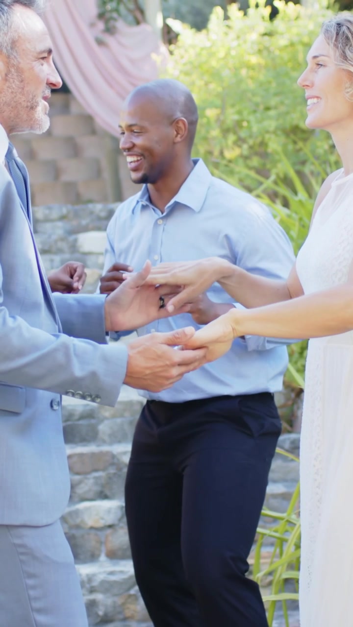 Coppia che balla, lui in abito grigio e lei in abito bianco, si tengono per mano in giardino mentre gli invitati applaudono, mostrando l'anello.