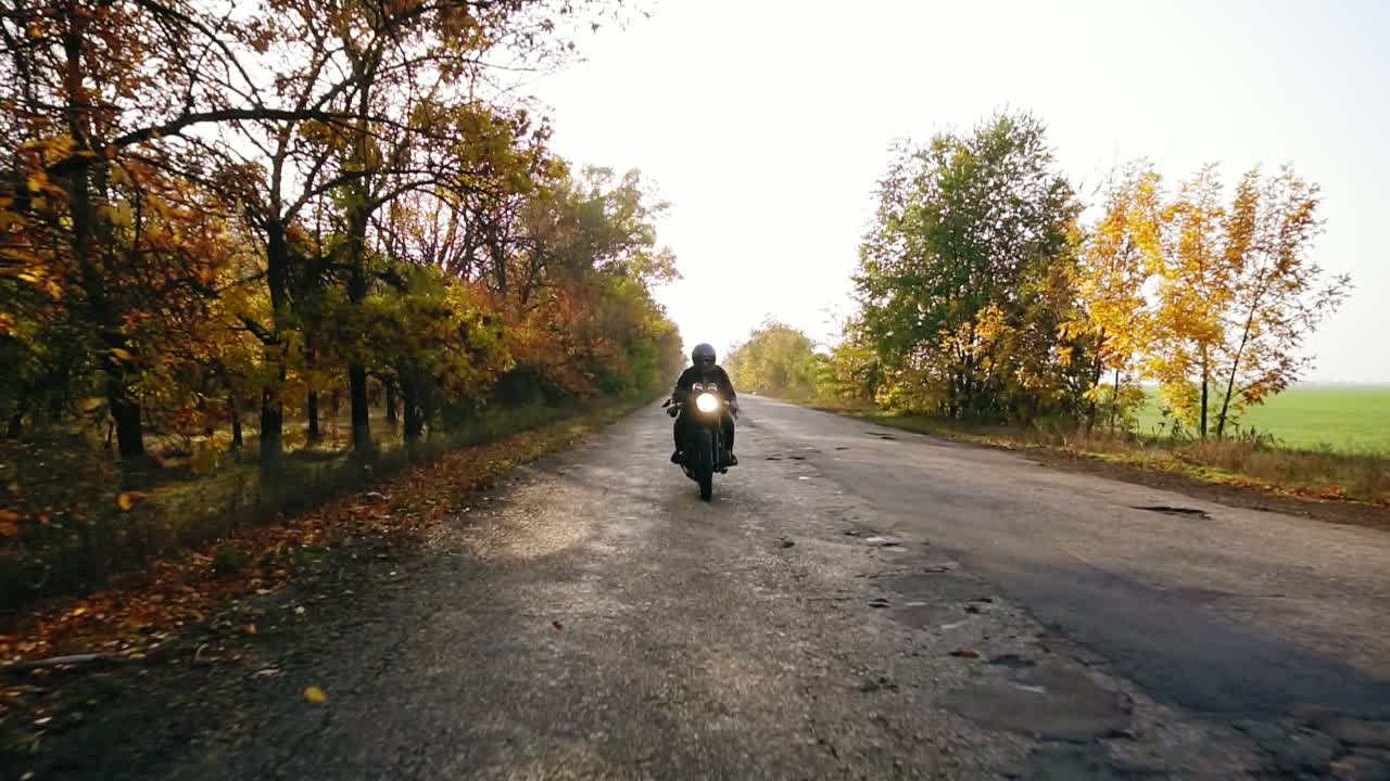 Unrecognizable man in black helmet and leather jacket approaching while riding a motorcycle on a road on a sunny day in autumn