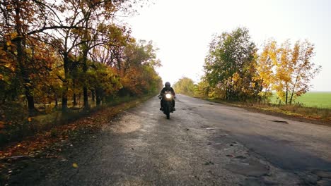 Unrecognizable-man-in-black-helmet-and-leather-jacket-approaching-while-riding-a-motorcycle-on-a-road-on-a-sunny-day-in-autumn