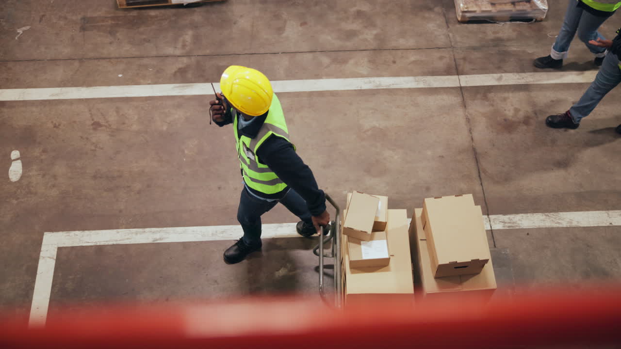 Warehouse Worker Pushing Hand Truck with Boxes and Talking on Walkie-Talkie