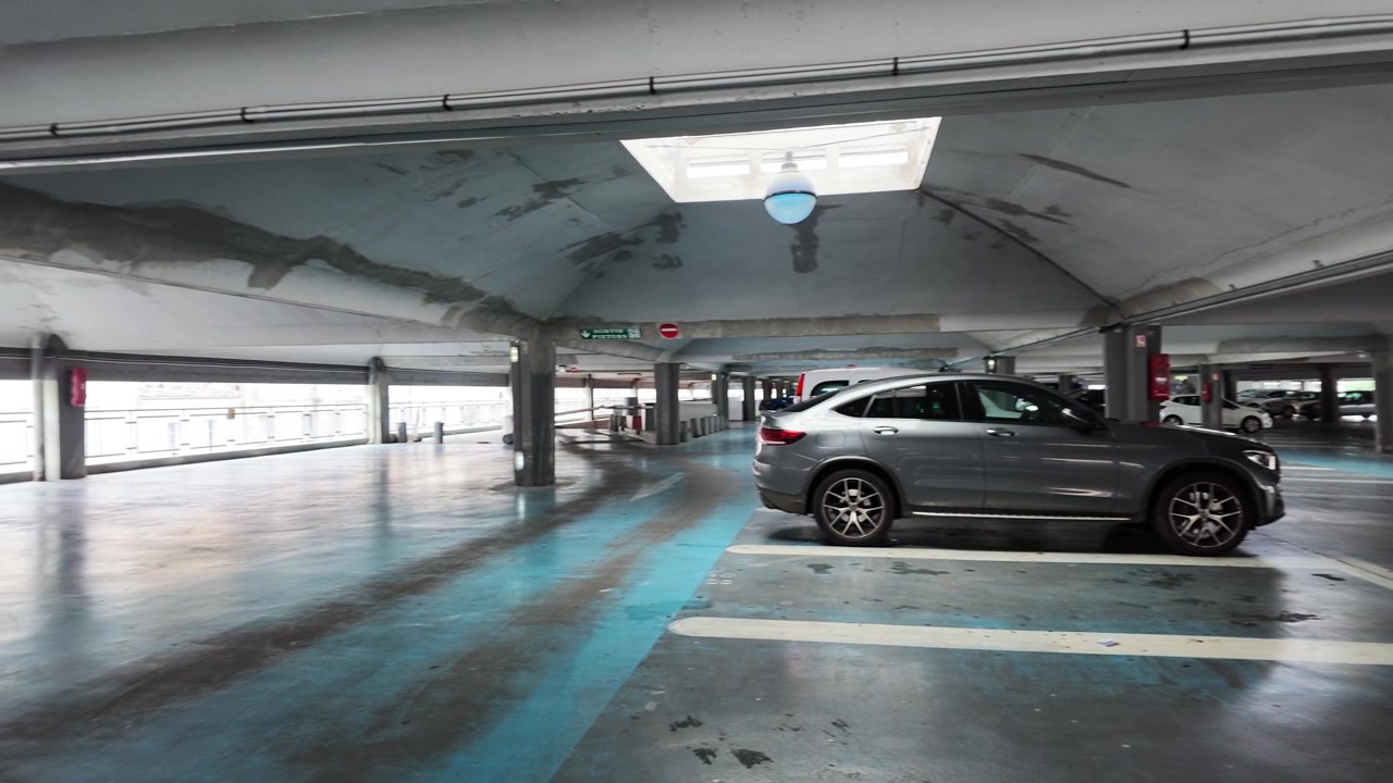 Plano geral estático de um SUV cinza manobrando em um estacionamento subterrâneo urbano.