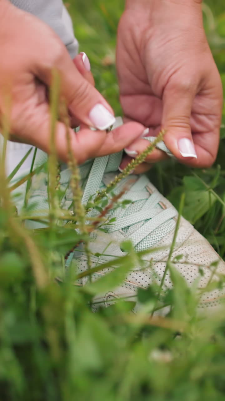 Primer plano de unas manos atando los cordones de unas zapatillas deportivas en la hierba. Zapatillas color menta y esmalte de uñas blanco, luz suave de la mañana, rocío sobre el trébol, preparación íntima para caminar o correr, enfoque delicado y ritual pausado.