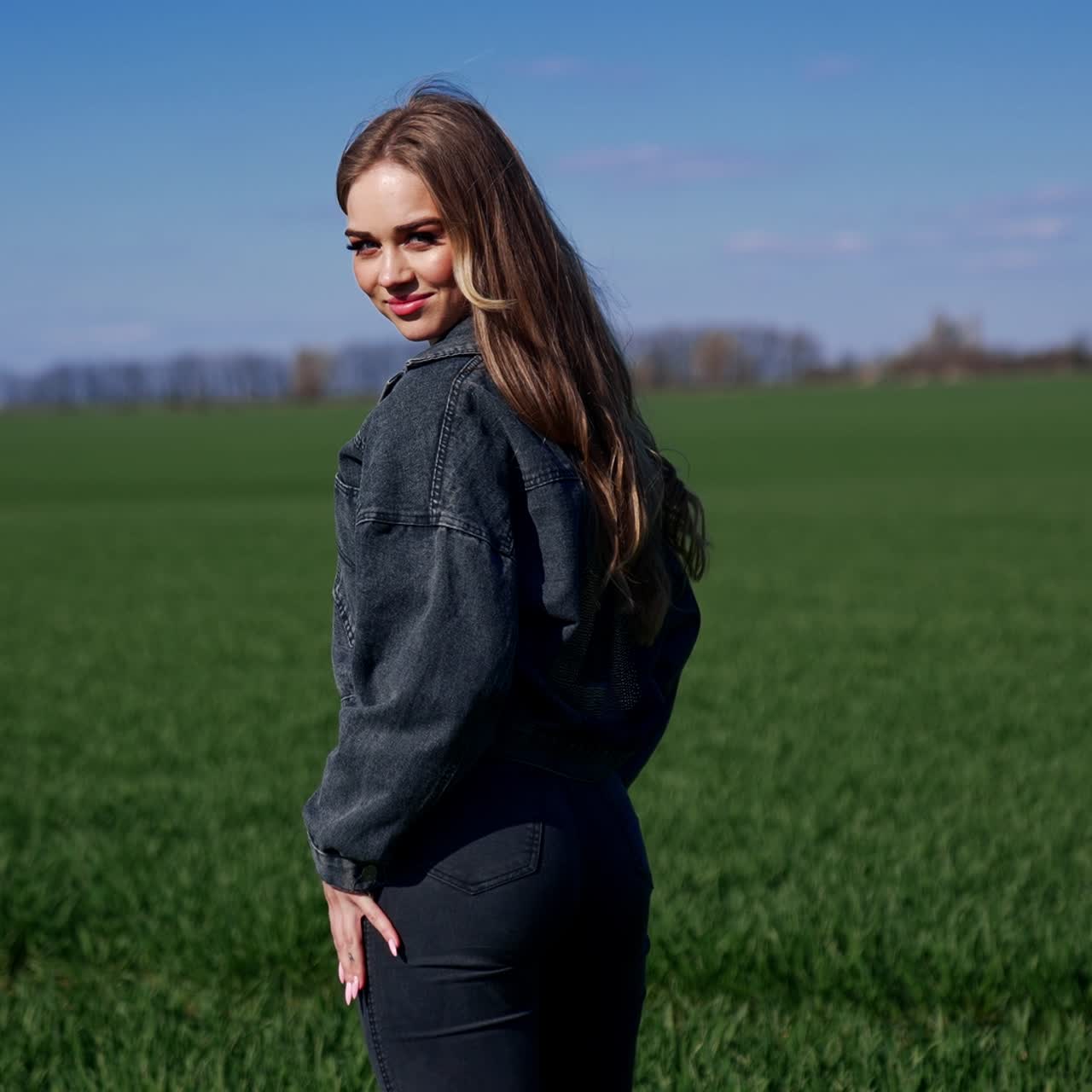 Ung kvinde udendørs. Attraktiv pige i denim-sæt på en grøn mark. Smuk pige med langt hår vender sig mod kameraet og smiler på en eng. Bagfra