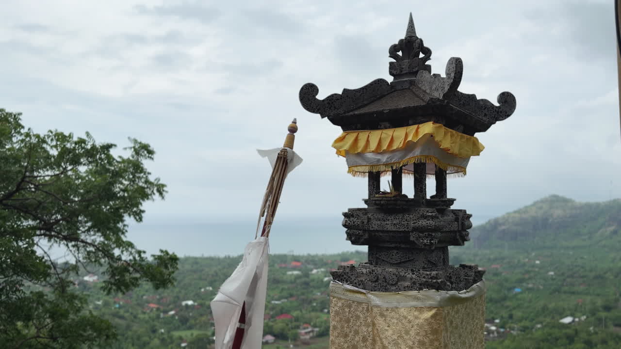 Balinese shrine overlooks Pemuteran village and coastline. Traditional stone lantern and ceremonial cloths create a serene spiritual atmosphere in tropical surroundings.