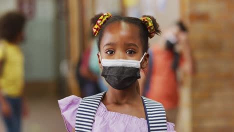 Portrait-of-african-american-schoolgirl-wearing-face-mask,-standing-in-corridor-looking-at-camera