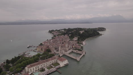 Toma-De-Drone-De-Simione-Italia-Mirando-La-Antigua-Fortaleza-En-Un-Día-Gris-Con-Barcos-En-El-Agua-Cerca-Del-Lago