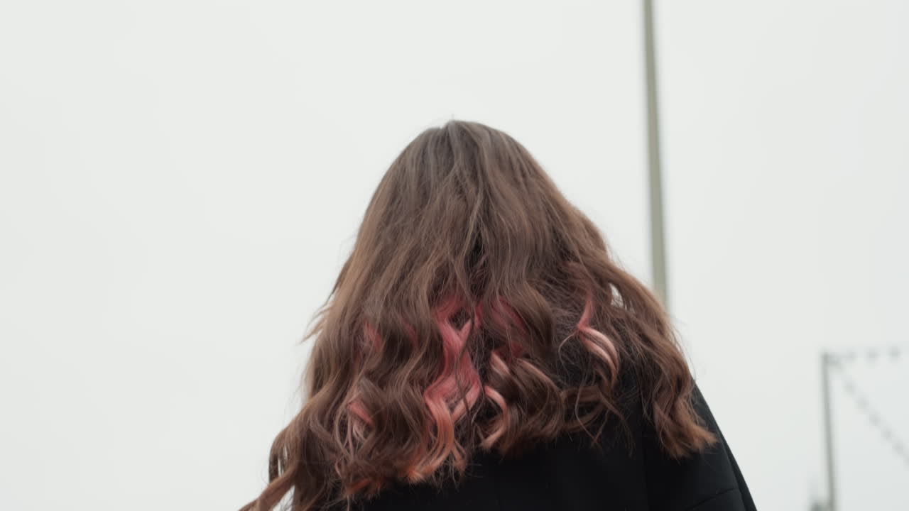 Giovane controluce che si sistema i capelli all'aperto. Figura in silhouette che cammina lungo una strada cittadina sotto un cielo nuvoloso. Un sentiero cittadino cattura uno studente introspettivo con capelli spettinati dal vento e punte colorate vivaci.