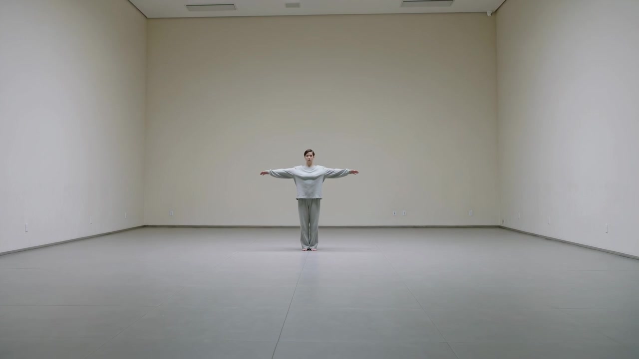 Dopo una pausa, una donna alza lentamente le braccia in posizione a T in una galleria, indossando un top chiaro e occhiali, mentre prova.