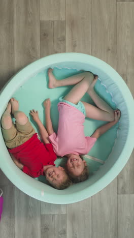 mom opens large sack with balls while happy children lying in pool. toddler boy and preschooler girl look forward for funny activity upper view