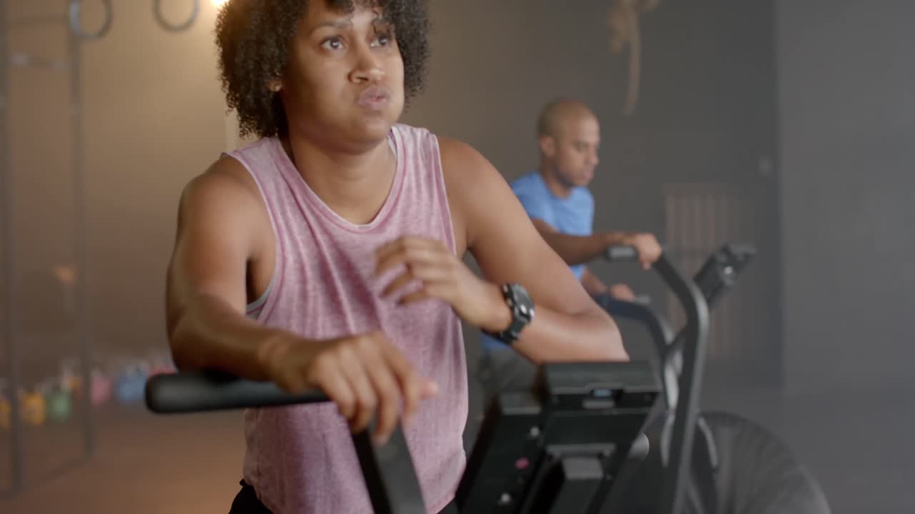 Donna afroamericana stanca in una lezione di fitness di gruppo eterogeneo che si allena su una cyclette ellittica, al rallentatore