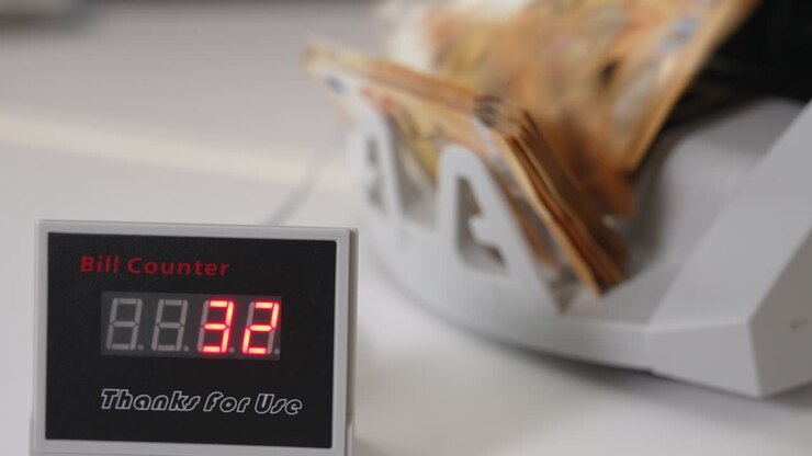 Orange banknotes falling on the holder of money counting machine. Male hand takes a wad of cash from equipment. Little screen showing the number of bills. Close up. Blurred backdrop.