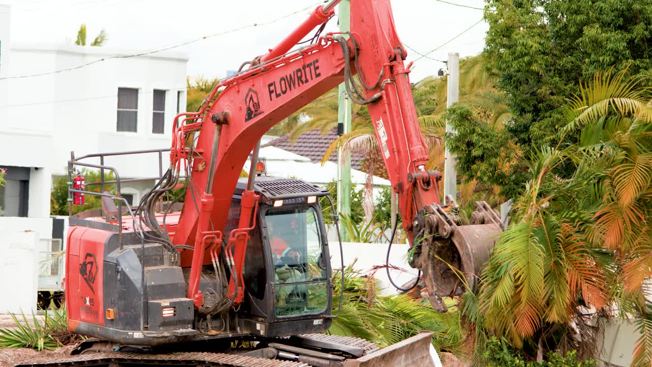 A red excavator removes a mature palm tree from a residential area in Gold Coast, Australia, under bright daylight with steady camera framing
