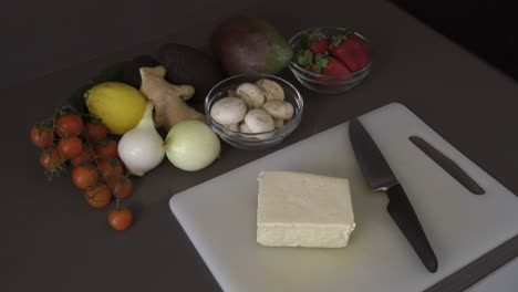 block of tofu and knife on the chopping board with various kinds of fruits and vegetables on the side. - high angle shot