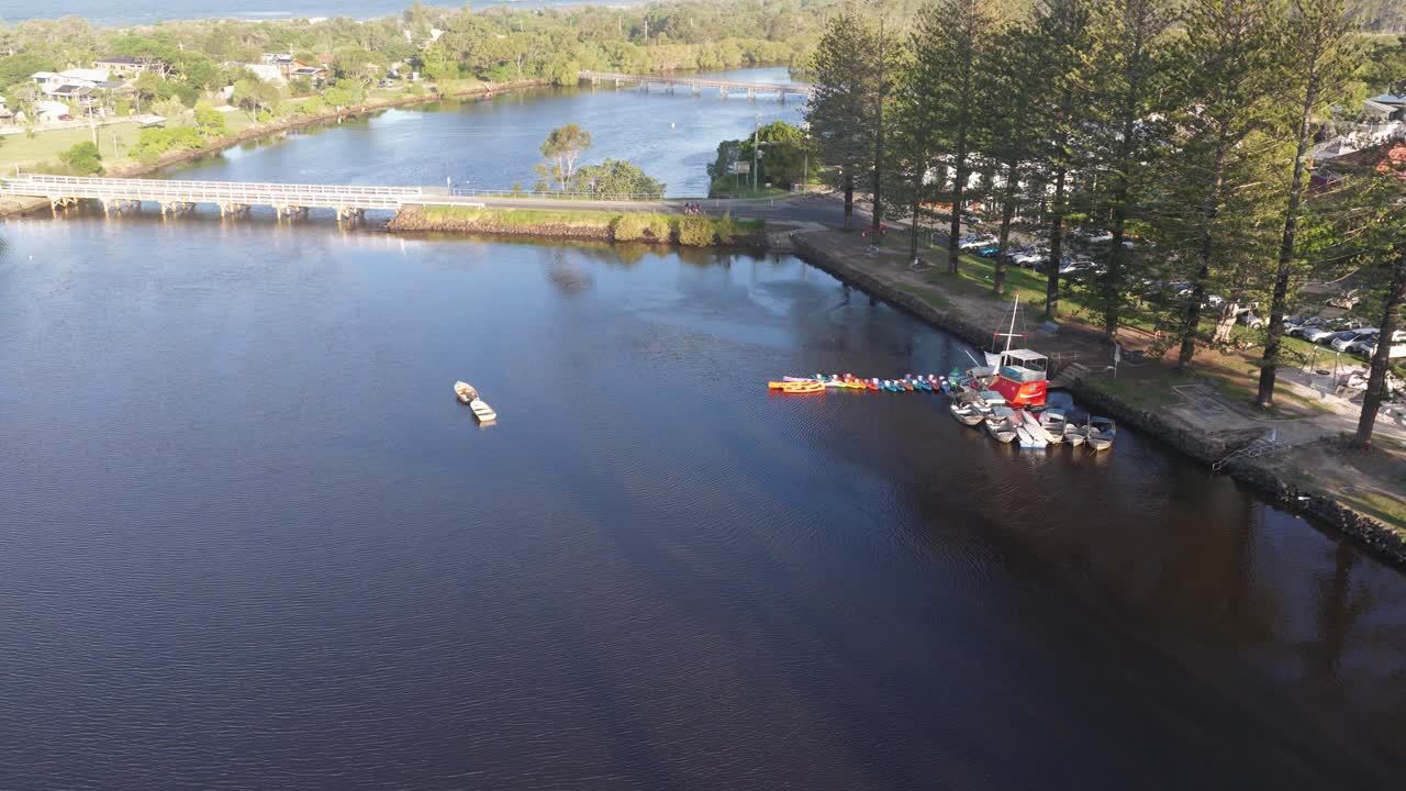 Aerial footage captures boats on a serene river surrounded by lush greenery and a clear sky in Brunswick Heads