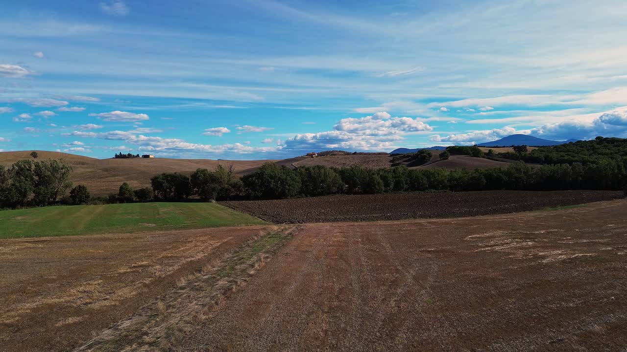 Aerial view of vast Tuscan fields with rolling hills, patches of greenery, and a vibrant blue sky with scattered clouds.