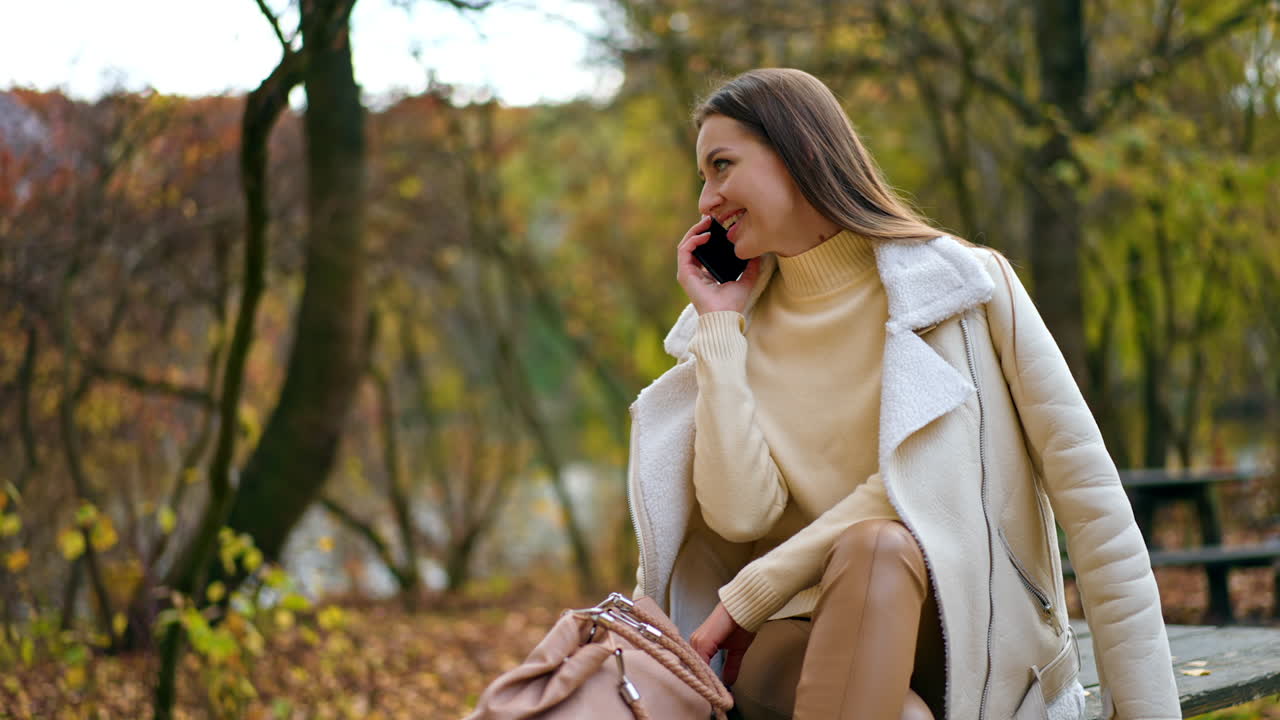 Happy smiling lady having phone conversation outdoors. Positive woman talks and smiles sitting on the old wooden table in the park in autumn.